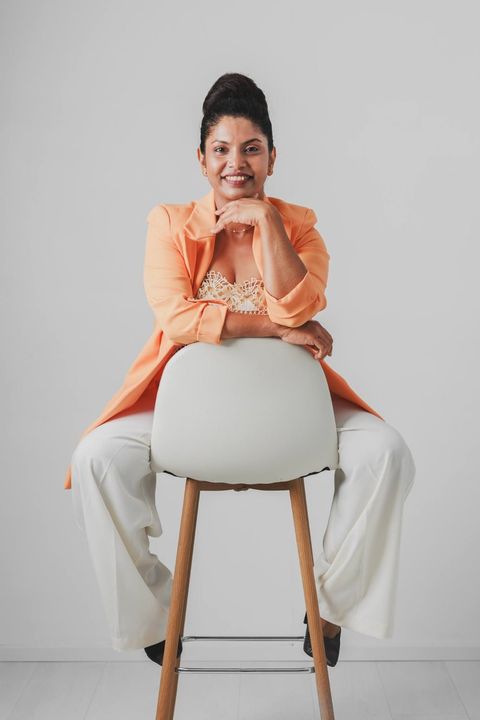 Thenu, fashion designer in Tampere, smiling on a studio stool in a peach blazer and white trousers